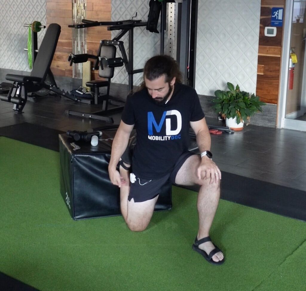 Dr. John with his right knee on the ground and his right foot ontop of a workout box. His left foot is on the ground in a 90 degree angle. He is stretching his right quad and tendon